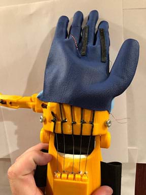 A blue rubber glove with black sensors on the back that have wires out of either end. The glove is on a 3D printed prosthetic hand with the fingers extended. 