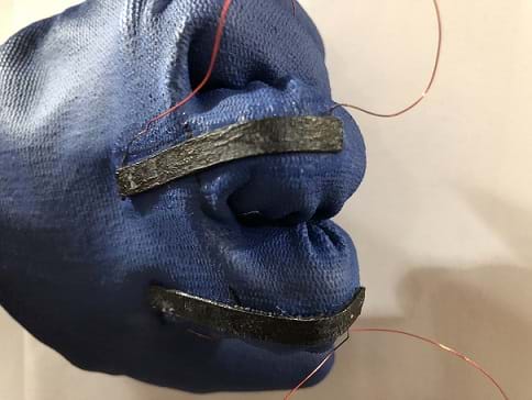 A blue rubber glove with black sensors on the back that have wires out of either end. The hand inside the glove is clenched in a fist.