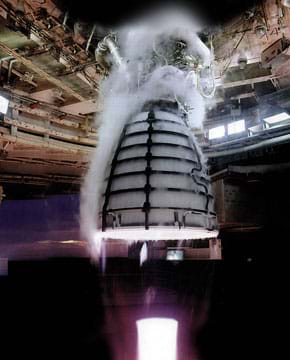 A photograph shows an engine undergoing a hot-fire test. The engine looks like a half of a 3D ellipse covered in a grid-like casing and attached at its top to a structure designed for testing. Surrounding and above the engine is a cloud-like substance, which is the condensation produced from the propellant.