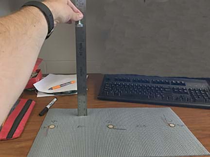 A photograph shows a person’s arm and hand holding a bolt 25 cm above a rubber mat (the same one shown/described in Figure 1) on a tabletop. 