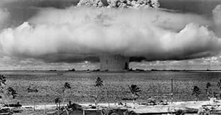 A black and white photograph from an island shows a mushroom-shaped explosion above the water at sea on the far horizon. This shows a nuclear weapon test by the U.S. military at Bikini Atoll, Micronesia, on July 25, 1946.