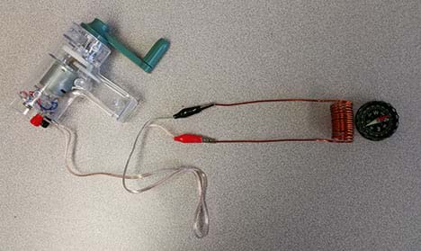 A photograph shows a compass sitting next to a coil of copper with one end of the coil’s copper wire connected to a red alligator clip and the other end connected to a black alligator clip. In turn, the clips are connected to wire from a small device that looks like a clear plastic gun with a spinning crank at the butt end (a hand-held generator).