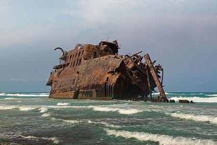 A large shipwreck is shown on choppy and wavy waters. The sky is blue and cloudy. The ship appears to be made out of metal, but is completely rusted with a brown/red color. The front of the ship is worn away and missing. One of the upper floors is angled and falling into the water. 