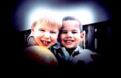 A photograph shows two smiling boys standing side-by-side, each holding a ball. The edges of the image are black, permitting only the center of the scene to be seen. 