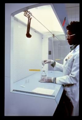 A photograph shows a lab tech in a white coat and gloves measuring liquid chemicals in a laboratory hooded area.