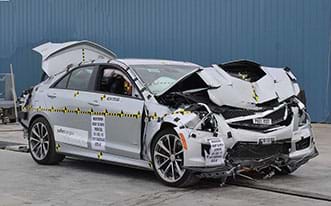 A photograph shows a silver sedan after a vehicle crash test, with a good view of the front and passenger sides of the vehicle. The hood, bumper, grill and engine are smashed and crumpled. The front passenger air bag and window air bag have been deployed. The trunk is open.
