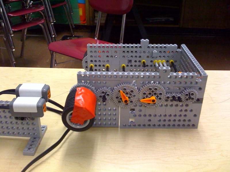A photograph shows a contraption made of LEGO parts. Two color sensors are focused on two wheels taped together that are attached to a large spur gear mounted on a three-sided LEGO wall structure. The gear is part of a gear train composed of eight gears of different sizes, positioned in a line.