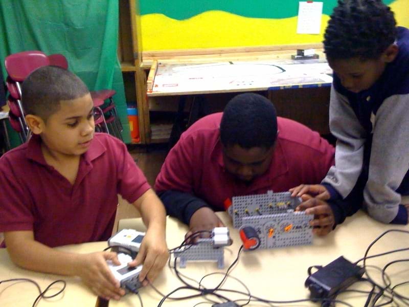 Three boys at a table set up their experiment by preparing the robot, fixing the gears and setting up the light sensor.