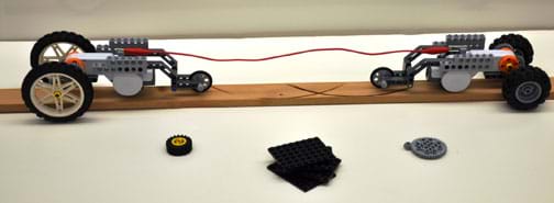 Photo shows two three-wheeled robots facing each other at opposite ends of a board, with a string between them, attached to each. In front are extra pieces, a wheel, a gear and some LEGO pieces. 
