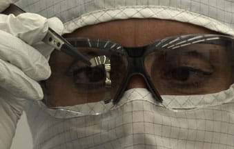A photograph shows a technician in a white clean suit and lab glasses inspecting what looks like a tiny and shiny, thin and flexible piece of material, held by tweezers.