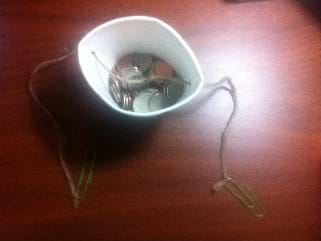 Photo shows a paper cup partially filled with coins. A string runs through two holes on opposite sides of the cup near the rim of the cup. Each end of the string is tied to a paper clip.