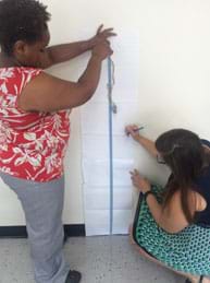 A photograph shows two female teachers testing the activity. One drops Washy tied to a few rubber bands while the other observes, measures and records the resulting displacement from the fall on paper taped to the wall.
