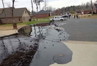 A photograph shows the black slick of an oil spill on a residential cul-de-sac street, spilling over a concrete driveway and contained in a pool by a curved concrete curb. The crude oil spill came from a rupture in ExxonMobil’s Pegasus 850-mile IL-to-TX pipeline in Mayflower, AR, in 2013. 