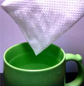 A photograph shows a folded paper towel with one corner just pulled out of a mug of water. Water from one tip of the paper towel has absorbed upwards to moisten a big area.