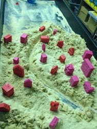A photograph shows a curving canyon (river pathway) cut through a mass of sand on which a scattering of 20+ miniature pink and red buildings are placed.