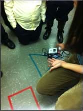 Several students and a teacher gather around a LEGO robot that is traveling the perimeter of a taped triangle on a linoleum floor using the robot's light sensor and programming that directs the robot to follow a colored line.