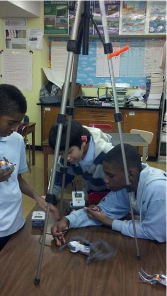 A photograph shows three boys working with LEGO bricks and servo motors as a smartphone spins under a camera tripod on a table.