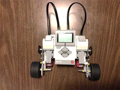 A photograph shows a LEGO EV3 robot composed of a brick-sized computer (a palm-size plastic device with buttons and display) with two servo motors, each attached to a three-gear drive train and two beefy 4x4 wheels.