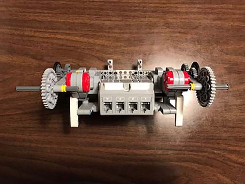 A photograph shows a LEGO EV3 robot—a shoebox-sized device made of plastic components. The view shows the four-port side of a LEGO EV3 brick with two attached servo motors, each attached to multi-gear drive trains with axles (no wheels are attached in this view).