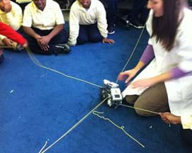 Three students and a teacher observe the collision of two LEGO robots where two lines of yarn cross on a carpeted floor.