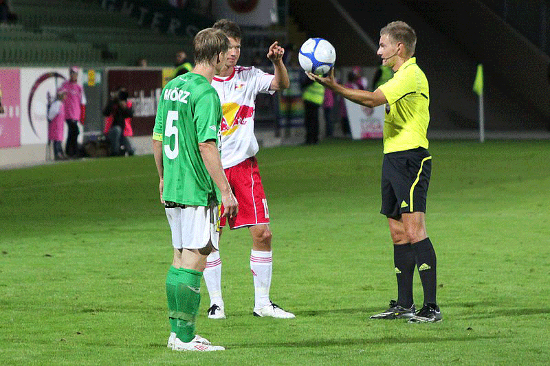 A photograph shows soccer referee standing next to two soccer players, about to drop the soccer ball.
