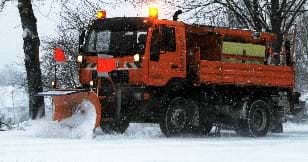An image of a large orange truck with a snowplow attached to the front, pushing snow along a tree-lined road.
