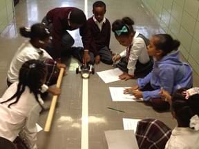 A photograph showing seven students in a semi-circle around the robot testing station. A robot pushes a container of sand along the length of track.