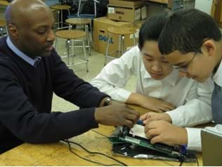 Photo shows two students constructing a traffic light circuit on a breadboard. An adult is assisting with the construction of the board.