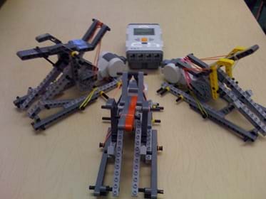 Photo shows three tabletop-sized devices made from LEGO pieces and rubber bands, with a LEGO brick sitting nearby.