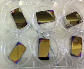 A photograph shows six silicon wafers coated with a doped polystyrene film. The rectangular pieces are mostly a reflective brass color with shades of orange, red, blue and purple at the corners and edges. The color variations indicate thickness variation.