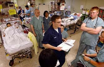 A photograph in an open hospital ward with many patients in beds and many medical workers and a clinical engineer conducting a meeting.