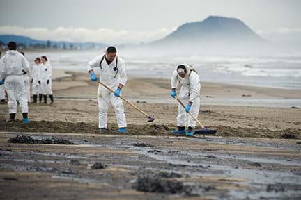 A photograph shows six people in white coveralls using rakes and hoes to clean Papamoa Beach in New Zealand after an oil spill reached the sandy shore from the grounded ship Rena.