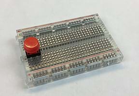 A photograph shows a breadboard with a red pushbutton placed so that one pair of its legs is on either side of the breadboard gap.