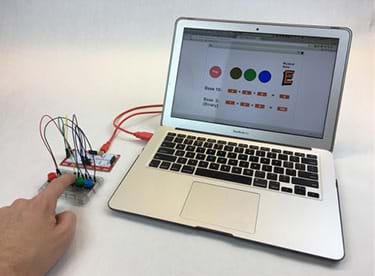 A photograph shows a laptop connected by a USB cable to a MaKey MaKey that is wired to four colored pushbuttons. A finger presses one button. On the screen is the binary trumpet program.