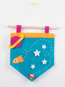 A photograph shows a finished light-up pennant made from felt that is hanging from a wooden dowel and string with a push-pin into the wall. On the pennant, multi-color felt cutouts depict a space scene with planets, stars and a rocket.