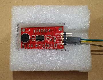 A photograph shows a small red sound detector (circuit board) with header pins connected using female-to-female jumper wires, nestled into a rectangular cut-out in a piece of white packing foam.