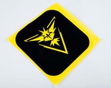 A photograph shows a square black iron-on fabric patch lying on a piece of yellow felt. Cut-outs in the patch let the yellow felt show through, revealing a Pokémon Zapdos species graphic that looks like a stylized bird with spread wings and spiky tail and head feathers. 