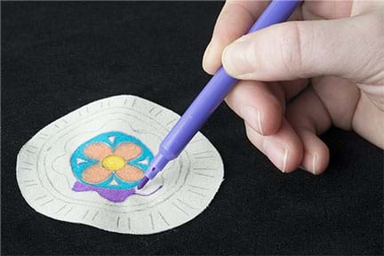 A photograph shows a hand holding a fine-tipped purple marker and coloring in a flower design on white fabric, which is the front of the pin.