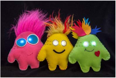 A photograph shows three plump “plush” toys made of felt, each embedded with a LilyPad microcontroller and two LEDs as glowing eyes. The monster blob shapes loosely indicate arms and legs, with faux fur and felt feather shapes sewn into the top seams as hair.    
