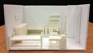 A photograph shows a diorama-type scale model of a dorm room that looks like a box with the top and one long rectangular sides open. Inside the “box” is assorted 3D-printed furniture along the walls (two beds, two desks, two chairs, two dressers, two bookshelves, and a few other organizational items) arranged in an open floor plan.