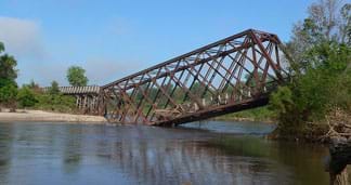 A photograph shows a collapsed steel truss bridge with one end sinking into the middle of a small river.