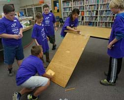 A photograph shows six youngsters in a classroom clustered around a board ramp from the table to the floor. One girl uses a wooden skewer to push a white Ping Pong ball down the ramp while a boy kneeling near the ramp bottom holds an envelope to catch the ball.
