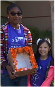 A photograph shows two girls wearing Elementary MESA Day shirts standing next to each other, looking at the camera. One holds her egg catcher made of cotton balls and crumpled tissue paper lining the inside of a cardboard box.