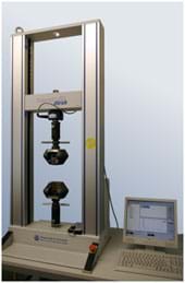 A photograph shows a desktop machine shaped like the frame of a tall open window with two sensors positioned from above and below that squeeze material placed between them. The force of the squeeze and the response of the material are recorded by a computer nearby.