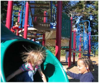 A photograph shows two young girls at a playground. One has just come down a plastic tube slide, and due to static electricity, her fine blond hair stands up in all directions around her head.