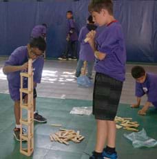 A photograph shows two boys using long, rectangular-shaped blocks as stacked columns and beams to make a tower on the floor. One boy is observing the other as he carefully places a block.