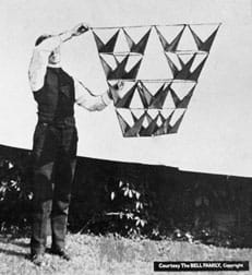 A black and white photograph shows a man using two hands to hold up and away from his body a 16-celled tetrahedron kite. The design includes opposing triangular vanes and was photographed outside against a white sheet.