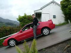 A photograph shows a man standing next to a sports car that is parked on a very steep hill.