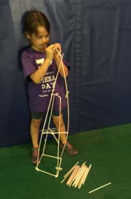 A photograph shows a young girl using plastic drinking straws and tape to build a tower structure that is almost as tall as she is.