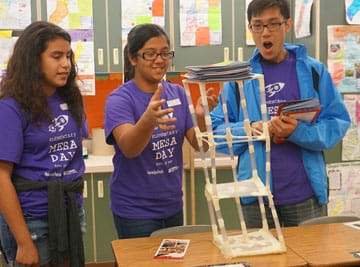 A photograph shows three students as they are adding magazines to the top of a 3-foot-tall straw tower on a desk to see how much weight it can hold.